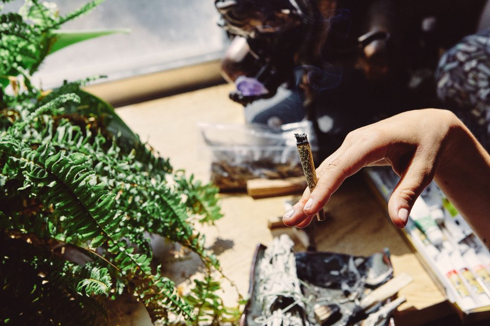 Hand holding a hand-rolled cigarette beside a fern and smouldering herbs