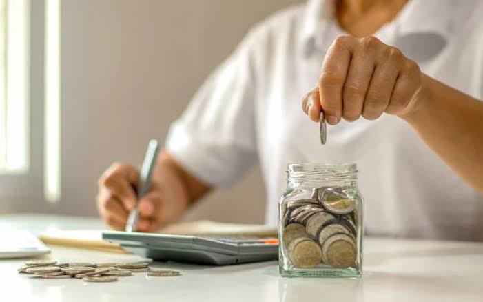 Hand dropping a coin into a jar beside a calculator and notebook
